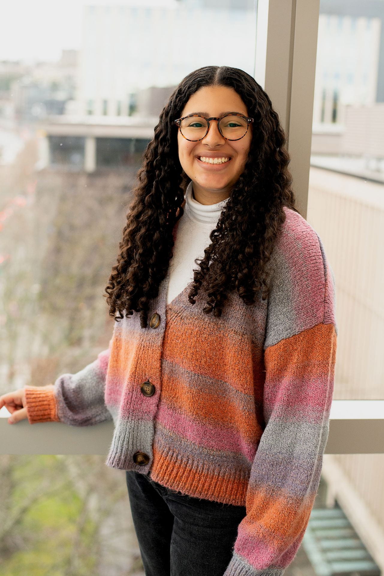 A woman with glasses and long, dark brown, curly hair, wearing a colorful sweater.