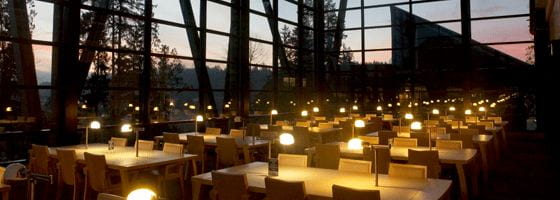Room dimly lit by table lamps, floor to ceiling windows along every wall with sunset in the background, and many wooden group tables and wooden chairs.