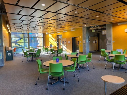 Building lobby with several circular tables with four green chairs around each of them. Floor to ceiling windows along one wall of the lobby.