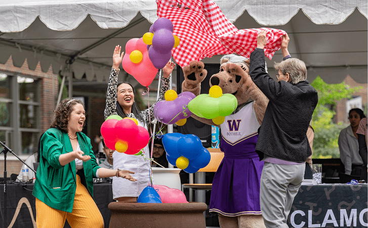 Three people and the UW Bothell husky mascot with their arms up releasing a string of flower balloons for a club event.
