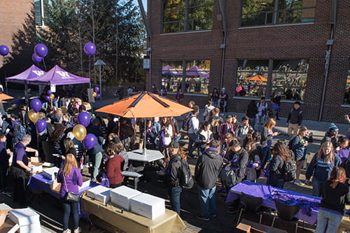Campus promenade outdoor event with purple and gold balloons and decorations. Many people standing and checking out the booths.