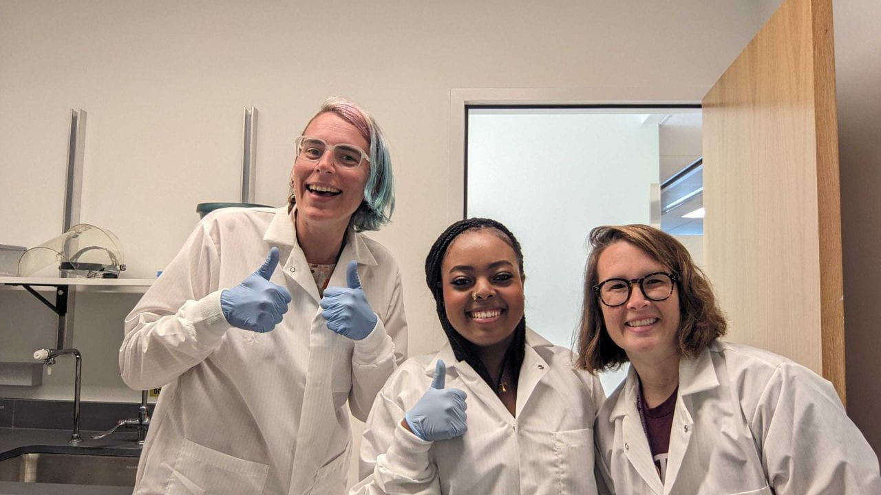 Valerie, Hanna, and Maddie are wearing lab coats and smiling, excited to pour their first gel!