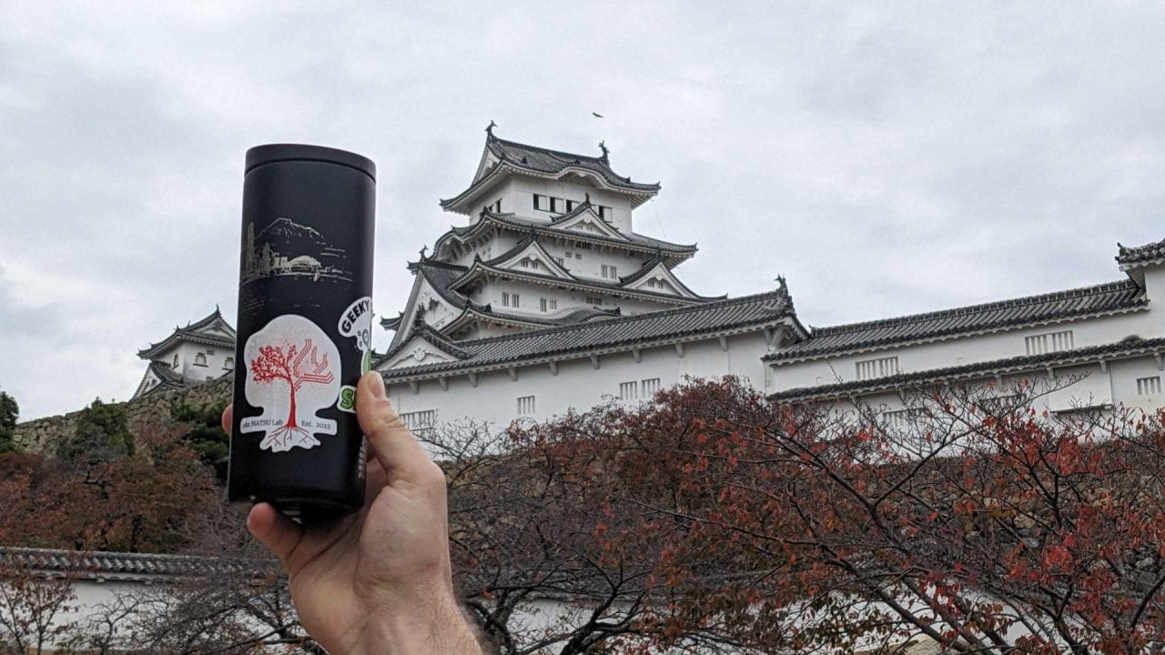 A water bottle with a Akamatsu-lab sticker is held up with Himeji Castle in the background. The Akamatsu lab was founded in 2022. Around 1300, Akamatsu Norimura built a temporary fort on the hill that would eventually become Himeji Castle.