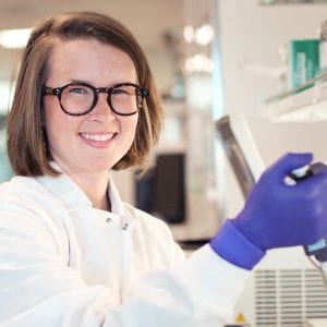 Maggie is wearing dark glasses and smiling at the camera. She is wearing a lab coat and gloves, and looks like she is pipetting.