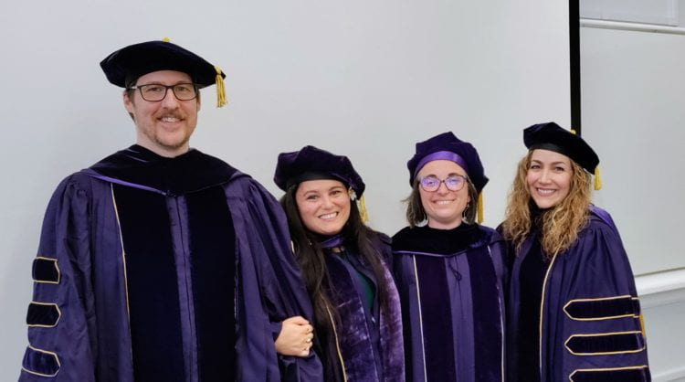 Four graduating PhD students stand next to each other while wearing black, purple, and gold academic regalia.