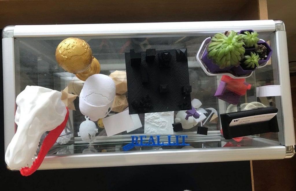 Top down view of a glass display case featuring 3D printed objects, including a skull, a globe, a planter, and a business card holder