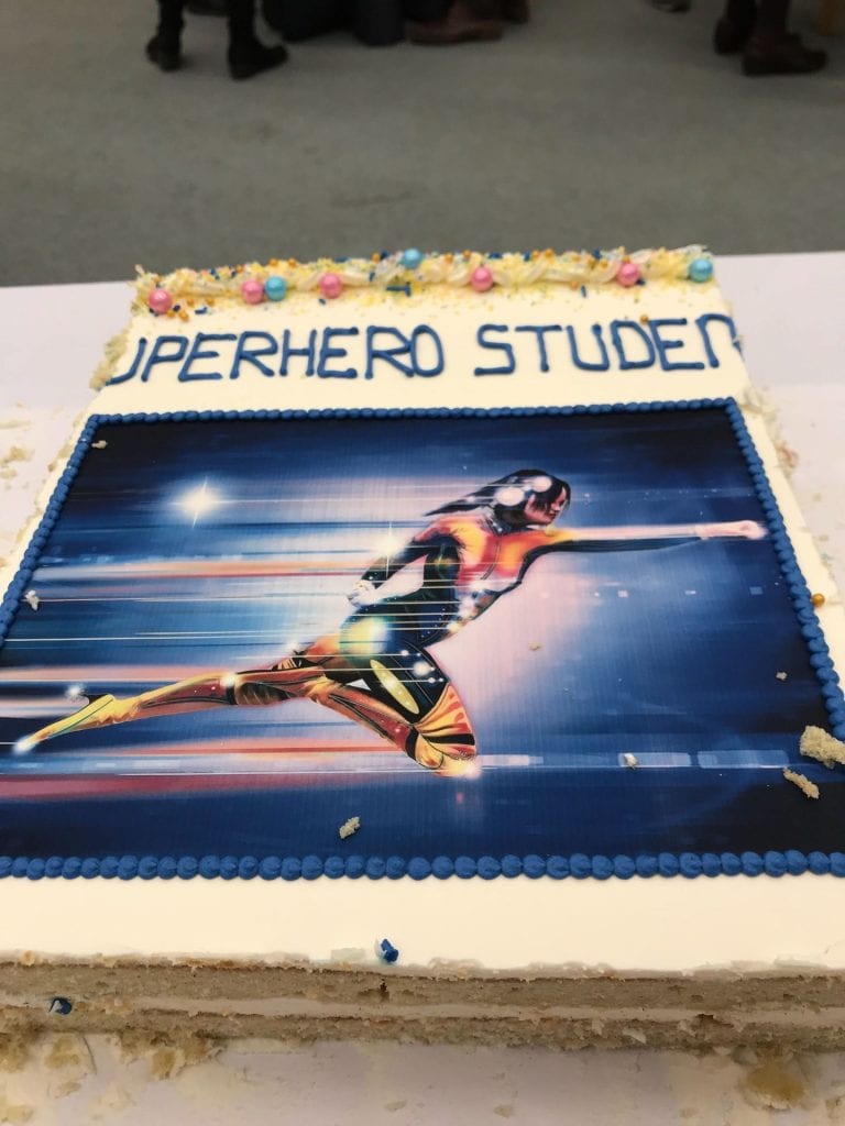photo of a sheet cake with "superhero student" written on top, and a picture of a female-bodied superhero