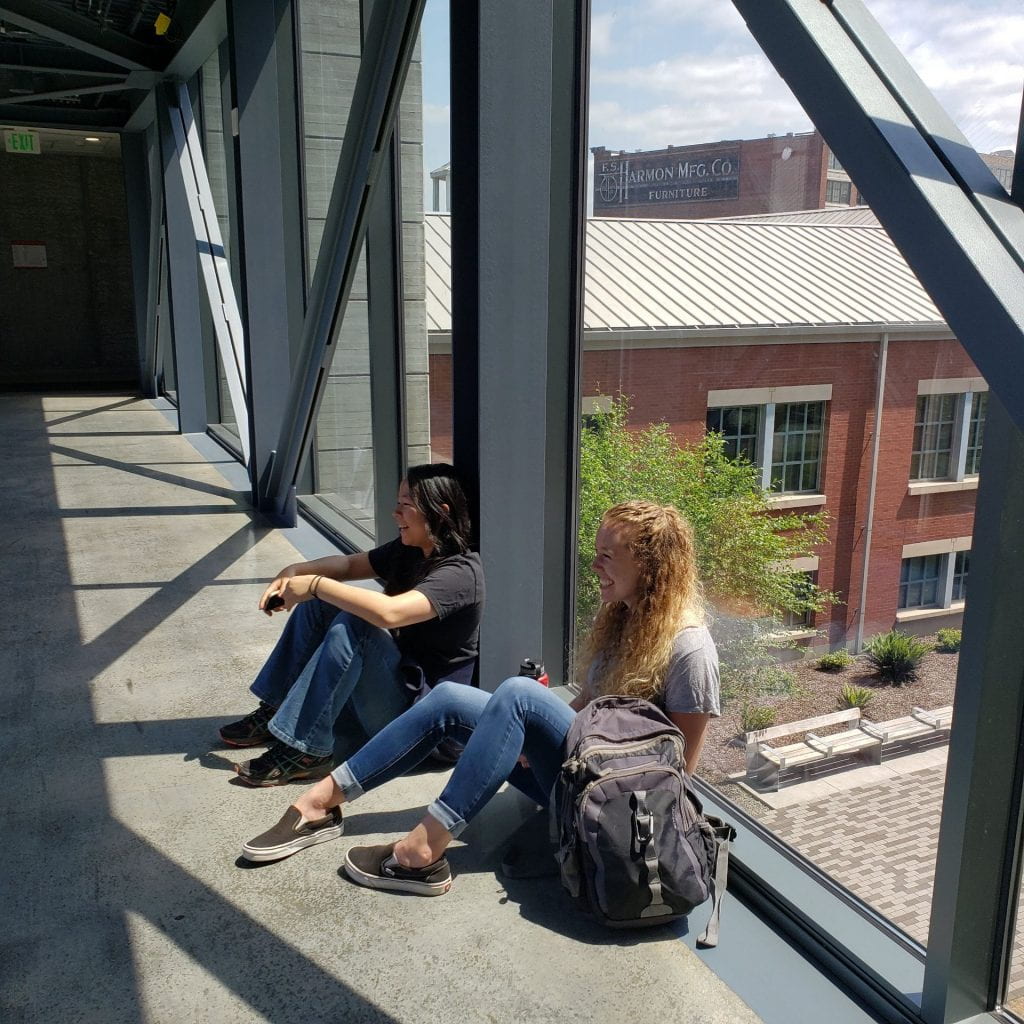 Two people sit with their backs against a glass wall a couple of stories above the ground behind them
