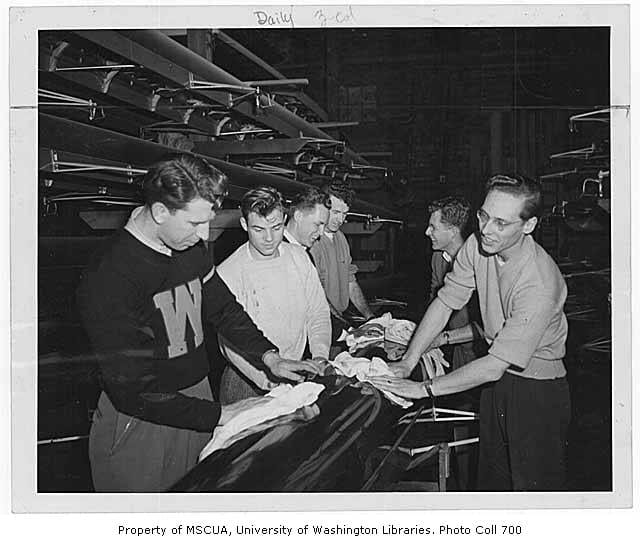 Black-and-white image of 6 people in a crew boat shed polishing the underside of a boat