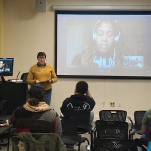 A person standing in front of a large projector screen where Angie Thomas is talking to the audience via Skype.