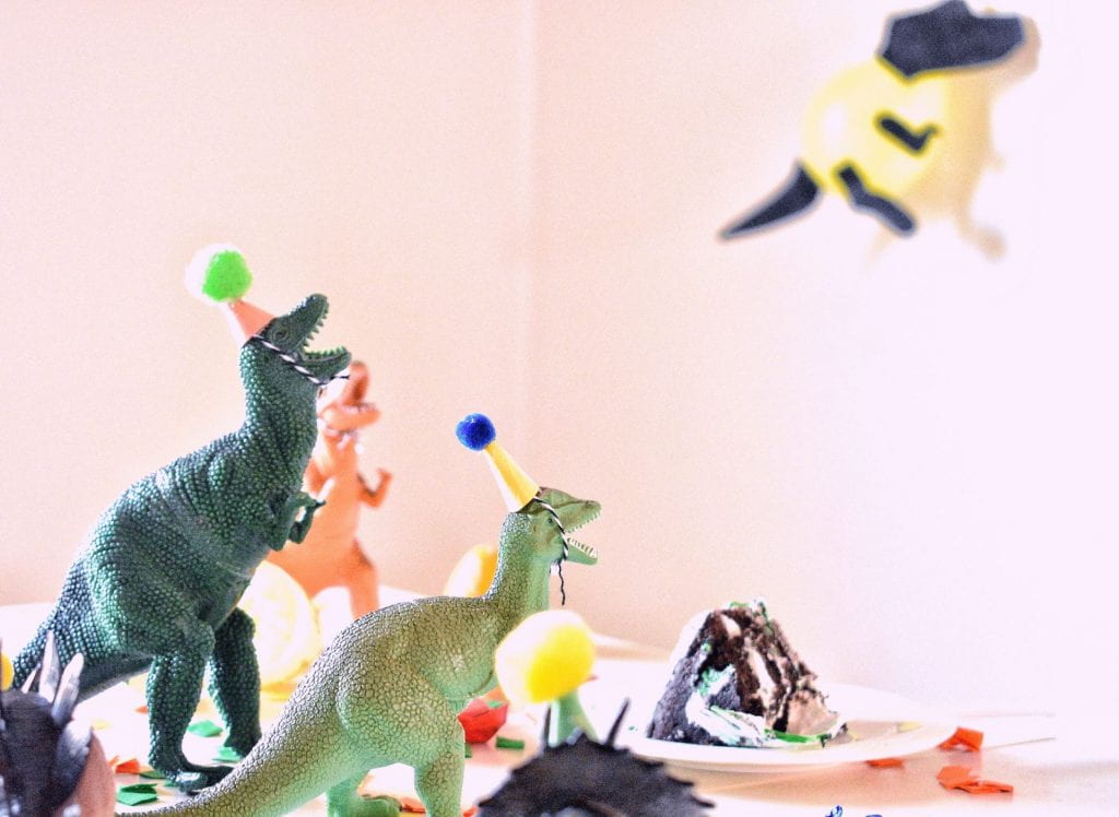 A group of toy dinosaurs wearing birthday party hats. In the background is a slice of cake.