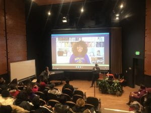 Image of Elizabeth Acevedo on a large screen in a University auditorium, participating in a skype call with students.