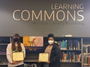 Two masked students in holding awards in front of a book shelf in a Library. Text reads: Learning Commons @ Tioga Library Building