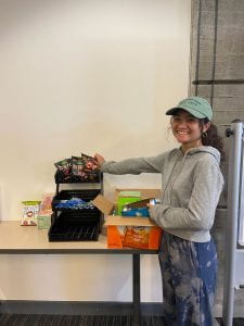 Young BIPOC woman with a baseball cap smiling into the camera, pointing at a selection of snack items being handed out for free in the library