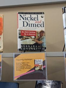 The cover of a book titled, Nickel and Dimed: On (Not) Getting By in America, by Barbara Ehrenreich. The cover features a white woman with curly blonde hair. She is wearing a blue shirt and is standing at a white counter, looking over her shoulder.The cover is accompanied by a shelf talker that reads: "Frequently Challenged: For being 'faddish,' 'of no moral value,' obscenity; promoting economic fallacies and socialist ideas, advocating the use of drugs, and belittling Christians."