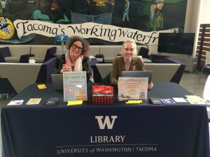 Two women sit at a table advertising the University of Washington Tacoma (UWT) Library Lab with signs, crackers, highlighters and paper flyers on the table in front of them